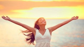 A woman, smiling with her arms wide open above her as she looks up to the sky full of hope and enthusiasm, the bright sun behind her and a body of water glistening and sparkling in the backdrop.
